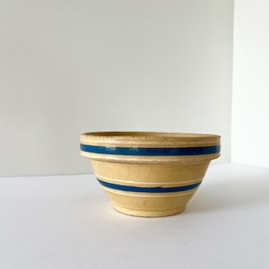 May include: A vintage yellow ceramic mixing bowl with two dark blue stripes. The bowl has a slightly flared shape and a simple, classic design. This bowl is perfect for baking and cooking.