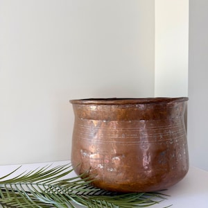 May include: A round, hammered copper pot with a wide opening and a slightly tapered base. The exterior surface features a weathered, metallic brown patina with visible horizontal etched lines encircling the upper portion of the vessel body.