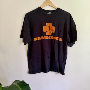 May include: A black short-sleeved t-shirt featuring an orange graphic logo on the chest. The design includes a stylized cross symbol above the text RAMMSTEIN in a bold, blocky font. The shirt is displayed on a wooden hanger against a plain wall.