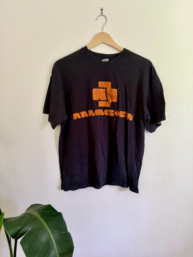May include: A black short-sleeved t-shirt featuring an orange graphic logo on the chest. The design includes a stylized cross symbol above the text RAMMSTEIN in a bold, blocky font. The shirt is displayed on a wooden hanger against a plain wall.