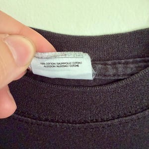 May include: A black t-shirt featuring a white fabric care label attached to the inner neckline. The label displays the text 100% COTTON / BAUMWOLLE / COTON / ALGODON / ALGODAO / COTONE, indicating the material composition of the garment.