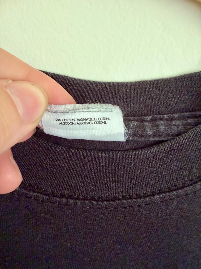 May include: A black t-shirt featuring a white fabric care label attached to the inner neckline. The label displays the text 100% COTTON / BAUMWOLLE / COTON / ALGODON / ALGODAO / COTONE, indicating the material composition of the garment.
