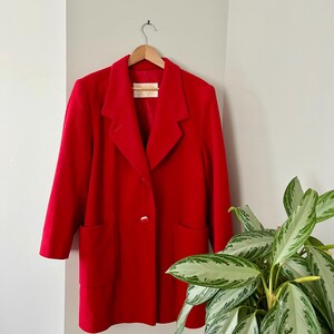 Op de afbeelding: Een felrode blazer met een klassiek ontwerp, met een reverskraag, twee voorzakken en een enkele gouden knoopsluiting. De jas hangt aan een houten hanger tegen een neutrale muur.