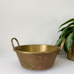 Puede incluir: Un cuenco de latón vintage con dos asas arqueadas. El cuenco tiene un diseño ancho y poco profundo con un borde ligeramente ensanchado. El latón tiene una pátina cálida y envejecida, con variaciones de color. Una pequeña olla de latón y una planta verde están en el fondo.