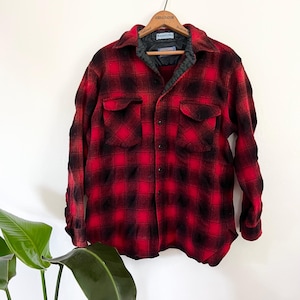 Może przedstawiać: Czerwono-czarna koszula w kratę z flaneli wisząca na drewnianym wieszaku. Koszula ma dwie kieszenie na piersi i pikowaną podszewkę. Na wieszaku napis "Ambassador" i etykieta "Western-Wear".