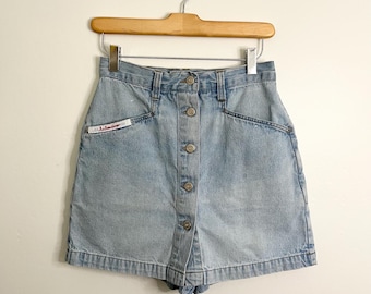 May include: Light blue denim shorts with a button-up front, pockets, and a small label with text. The shorts are hanging on a wooden hanger against a white background. The shorts have a high waist and a classic design.
