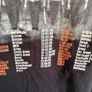 May include: A black t-shirt featuring a graphic of figures in white shirts and ties above a list of concert dates and venues from 2005, including locations like Brixton, Berlin, and various arenas printed in white and orange text on the fabric.