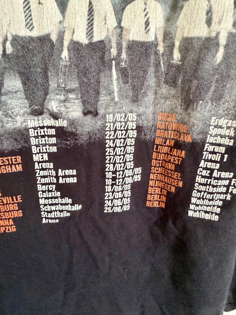 May include: A black t-shirt featuring a graphic of figures in white shirts and ties above a list of concert dates and venues from 2005, including locations like Brixton, Berlin, and various arenas printed in white and orange text on the fabric.
