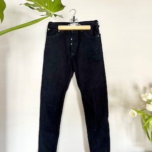 May include: Black denim jeans hanging on a wooden hanger. The jeans have a button fly and a classic five-pocket design. The dark wash of the denim creates a sleek, versatile look. The jeans are displayed against a white wall with a green plant and white flowers.