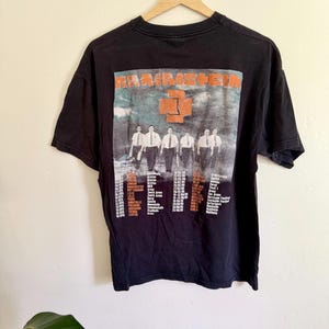 May include: A black short-sleeve t-shirt featuring a graphic print of the band Rammstein. The design includes the band name in orange, a central orange cross logo, a photograph of five individuals in white shirts and ties, and a list of tour dates.