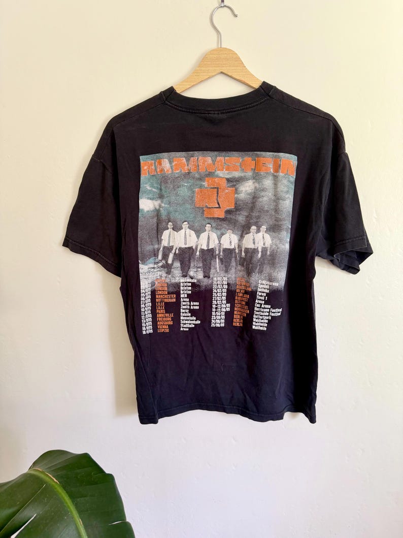 May include: A black short-sleeve t-shirt featuring a graphic print of the band Rammstein. The design includes the band name in orange, a central orange cross logo, a photograph of five individuals in white shirts and ties, and a list of tour dates.