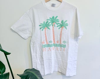 May include: White t-shirt with a tropical design. It features three palm trees in green and coral, the word "HAWAII" in coral, and green fish and waves. The shirt is displayed on a wooden hanger.
