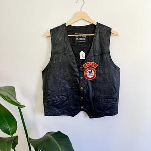 Może przedstawiać: Czarna skórzana kamizelka z dekoltem w serek i dwiema przednimi kieszeniami. Kamizelka ma złote guziki i czerwoną i białą naszywkę z napisem "RIDER". Kamizelka wisi na drewnianym wieszaku.