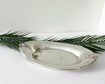 Può includere: Un vassoio ovale argentato vintage con decorazioni floreali sui manici. Il vassoio presenta un aspetto leggermente appannato, che suggerisce età e utilizzo. Sfondo bianco con una felce verde.