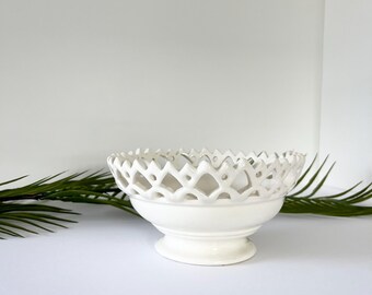 May include: A white ceramic bowl with a pedestal base and a decorative openwork rim. The bowl has a smooth, glossy finish and is set against a white background. The openwork design creates a delicate, airy aesthetic. The bowl is a decorative item.