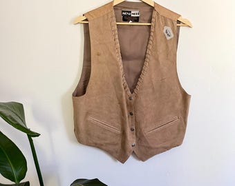 Puede incluir: Un chaleco de ante color canela con cierre de botones y dos bolsillos delanteros. El chaleco está colgado de una percha de madera contra una pared blanca. El chaleco tiene un diseño trenzado en los bordes y una etiqueta que dice "MONA LEE".