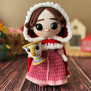May include: A crocheted doll wearing a red hooded cloak with white trim and a pink dress. The doll is holding a crocheted teacup with a face.