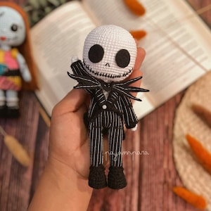 Puede incluir: Un muñeco de Jack Skellington de crochet que lleva un traje a rayas blanco y negro con una pajarita negra. El muñeco tiene la cara blanca con ojos negros y una boca cosida.