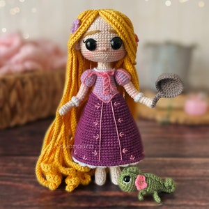 May include: A crocheted doll with long blonde hair wearing a purple dress and holding a frying pan. A green crocheted chameleon is sitting in front of the doll.