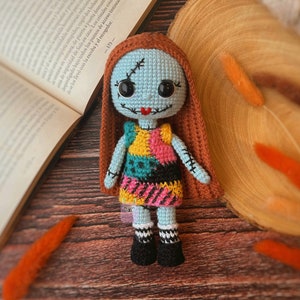 Puede incluir: Una muñeca de crochet con un vestido de retazos, cabello castaño y zapatos negros. La muñeca tiene una sonrisa cosida y grandes ojos negros. La muñeca está sentada sobre una superficie de madera con acentos naranja y marrón.