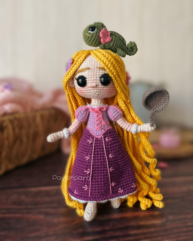 May include: A crocheted doll with long yellow hair, wearing a purple dress with pink accents. The doll is holding a brown spatula and has a green crocheted chameleon on its head. The doll is standing on a wooden surface.