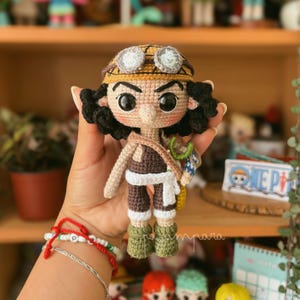 Könnte beinhalten: Handgefertigte Häkelpuppe einer Figur mit gebräuntem Gesicht, schwarzen Locken und großen schwarzen Augen. Die Puppe trägt einen beigefarbenen Hut mit Brille, ein braunes Outfit und grüne Stiefel. Sie hat eine kleine Tasche.