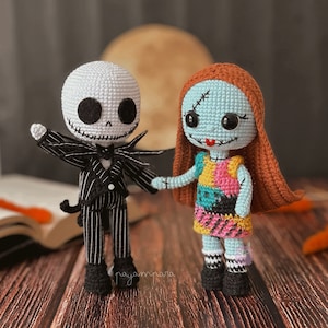 Pode incluir: Bonecas amigurumi de crochê de Jack Skellington e Sally do filme O Estranho Mundo de Jack. Jack está vestindo um terno listrado preto e branco com uma gravata borboleta. Sally está vestindo um vestido de patchwork colorido e tem cabelo ruivo. As bonecas estão em pé em uma superfície de madeira.