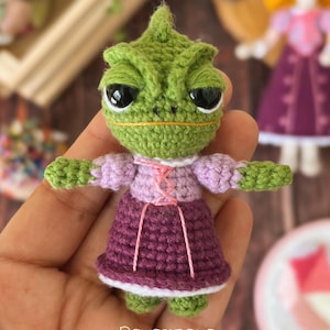 以下が含まれることがあります： ピンクのアクセントが付いた紫色のドレスを着た、緑色の編みぐるみカメレオン。カメレオンは大きな黒い目と小さなとがった鼻を持っています。