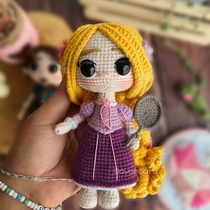 May include: Crocheted doll with long blonde hair, wearing a purple dress and holding a frying pan. The doll has large eyes and a sweet expression.