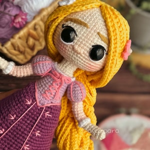 May include: A crocheted doll with long yellow hair, wearing a purple dress with pink accents. The doll has large brown eyes and a sweet expression.