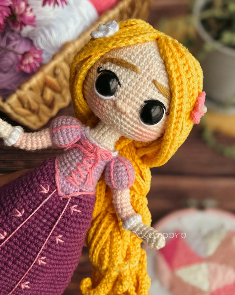 May include: A crocheted doll with long yellow hair, wearing a purple dress with pink accents. The doll has large brown eyes and a sweet expression.
