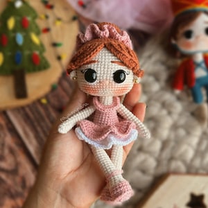 May include: A crocheted doll wearing a pink dress and a crown. The doll has brown hair, large eyes, and a sweet expression.