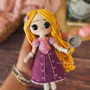 May include: A crocheted doll with long blonde hair, wearing a purple dress with pink accents and holding a grey pan. The doll has large black eyes and a sweet expression. The doll is handmade and has a charming, whimsical style.