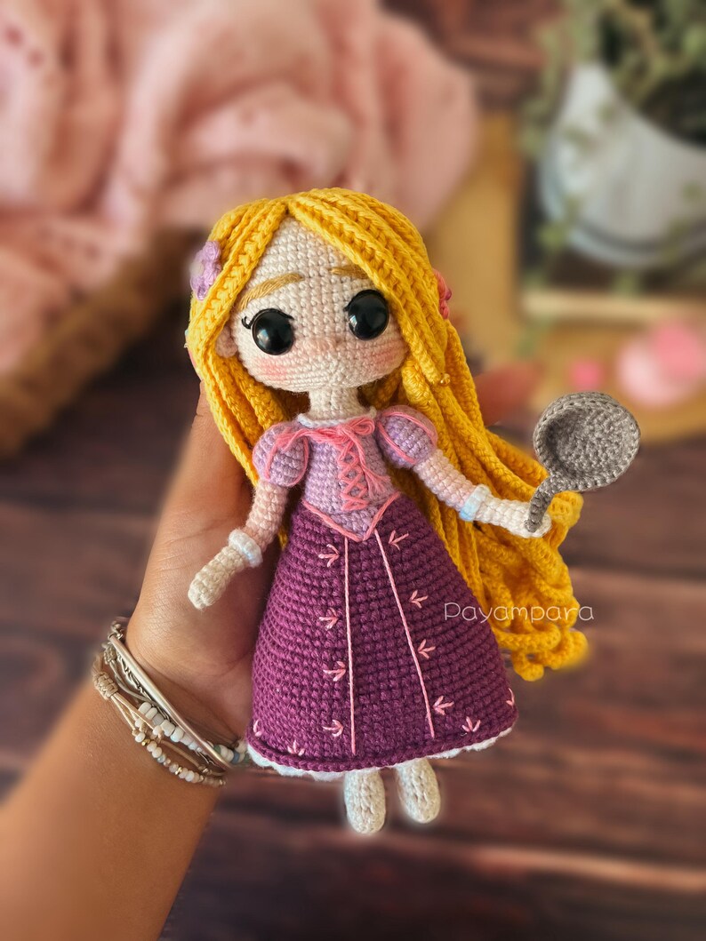May include: A crocheted doll with long blonde hair, wearing a purple dress with pink accents and holding a grey pan. The doll has large black eyes and a sweet expression. The doll is handmade and has a charming, whimsical style.