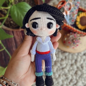 Puede incluir: Muñeca amigurumi de crochet con cabello negro, camisa blanca, cinturón rojo, pantalones azules y zapatos negros. La muñeca tiene una cara de dibujos animados con ojos grandes.