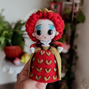Puede incluir: Muñeca de ganchillo de la Reina de Corazones de Alicia en el País de las Maravillas. La muñeca tiene un vestido rojo con detalles dorados, una peluca rizada roja y una corona dorada. La cara es blanca con sombra de ojos azul y ojos negros grandes.