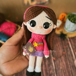 Puede incluir: Una muñeca de crochet de una niña con cabello castaño, que lleva un suéter rosa con un arcoíris y una estrella, sosteniendo un libro con una huella de mano en la portada.