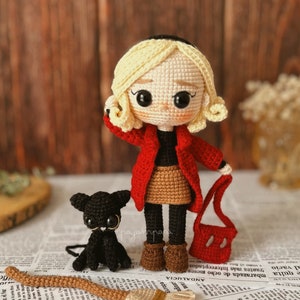 May include: Crocheted doll with blonde hair wearing a red coat, brown skirt, and brown boots. The doll is holding a red crocheted bag. A black crocheted cat sits next to the doll. A brown and beige crocheted broom is in the foreground.