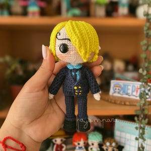 Puede incluir: Una muñeca de crochet con pelo amarillo, un traje azul y una camisa azul y blanca. La muñeca sostiene un pequeño objeto azul y blanco en su mano.