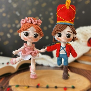 May include: A crocheted ballerina doll wearing a pink dress and a pink crown, holding hands with a crocheted nutcracker doll wearing a red and blue uniform and a red hat.