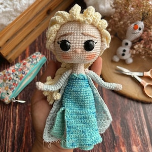 May include: A crocheted doll of Elsa from the film Frozen, wearing a blue and white dress with glitter. The doll has blonde hair and large black eyes.