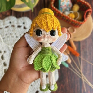 Puede incluir: Una muñeca de ganchillo de Campanilla con cabello amarillo, vestido verde y alas blancas. La muñeca sostiene una pequeña flor verde.