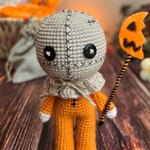 Puede incluir: Muñeco de ganchillo hecho a mano que se asemeja a Sam de la película Trick 'r Treat. El muñeco tiene una cabeza beige con detalles de costura negra, ojos de botón y un mono naranja. Sostiene una pequeña piruleta con forma de calabaza naranja.