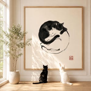 Könnte beinhalten: Ein quadratisches gerahmtes Wandbild mit einer Tuschezeichnung in Schwarz und Weiß, die zwei in einem Kreis zusammengerollte Katzen zeigt. Eine schwarze und eine weiße Katze sitzen auf dem Boden unter dem Bild und schauen nach oben.