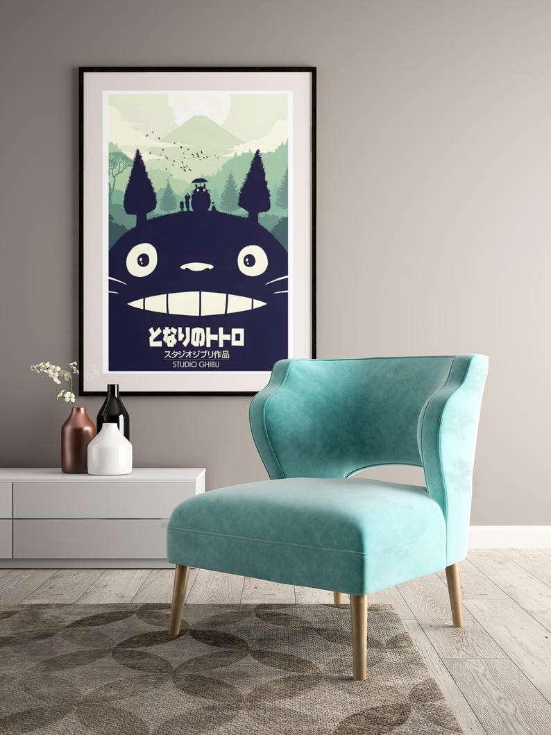 K&ouml;nnte beinhalten: A framed poster with a black frame, depicting Totoro from My Neighbor Totoro. The poster hangs above a teal armchair with wooden legs. The Japanese text is visible. The room also features a white cabinet and decorative vases.
