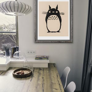 May include: A framed print featuring a black and white illustration of a Totoro character. The artwork is displayed on a wall above a wooden table. The room has a modern design with a window and a hanging light fixture.