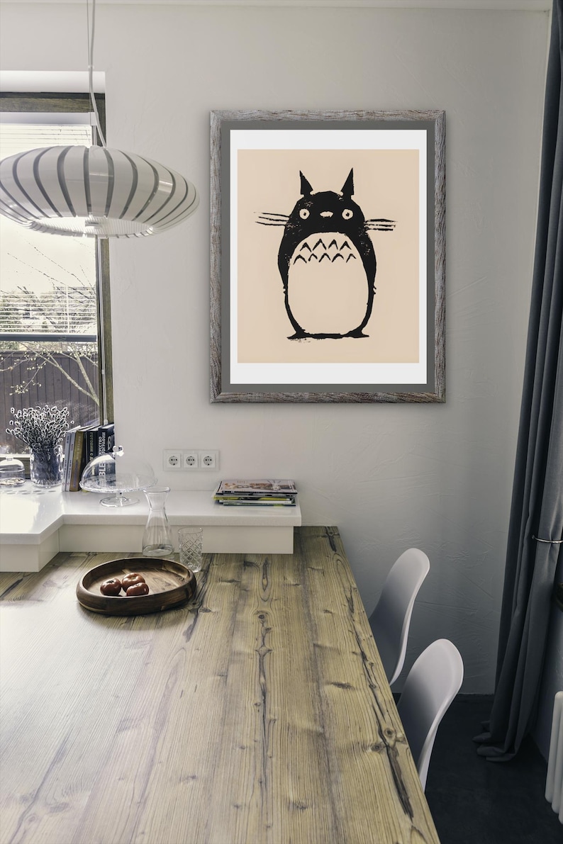 May include: A framed print featuring a black and white illustration of a Totoro character. The artwork is displayed on a wall above a wooden table. The room has a modern design with a window and a hanging light fixture.