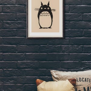 May include: Framed art print featuring a black and white illustration of a Totoro character. The artwork is set against a beige background and framed in a dark wood frame. The print is mounted on a dark brick wall, with decorative pillows in the foreground.