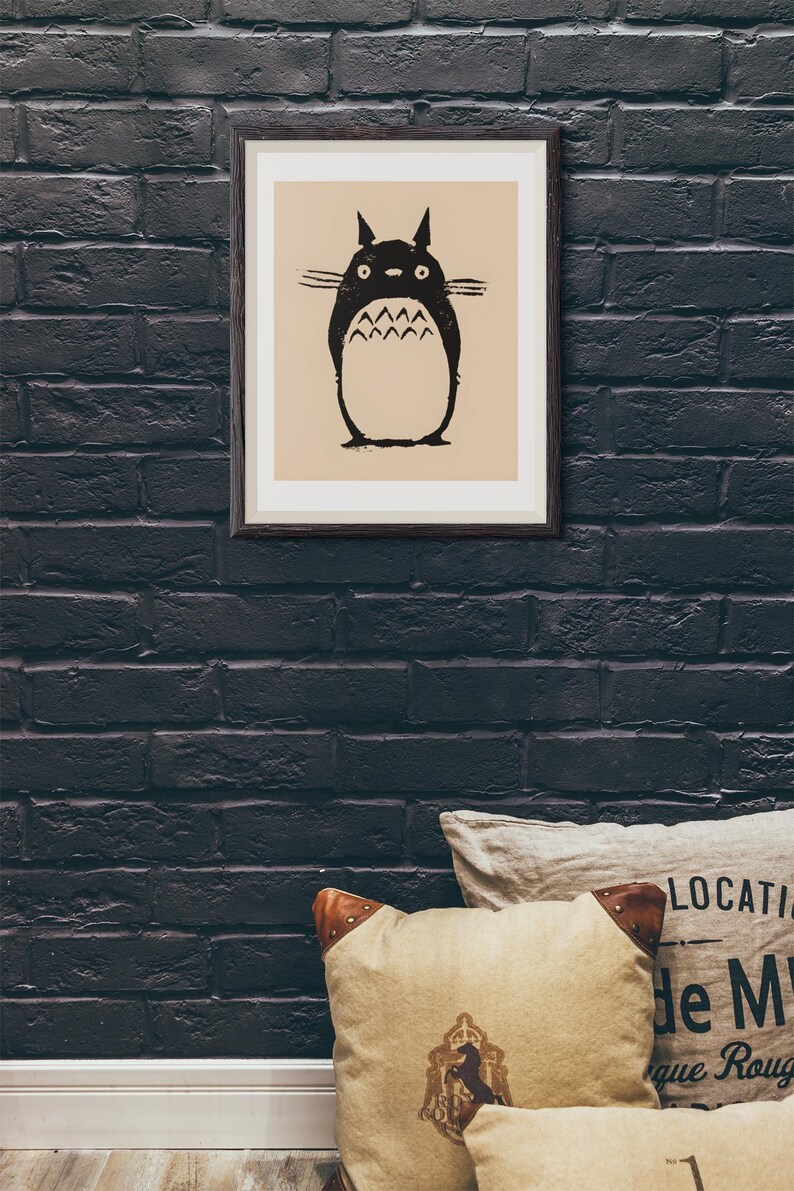 May include: Framed art print featuring a black and white illustration of a Totoro character. The artwork is set against a beige background and framed in a dark wood frame. The print is mounted on a dark brick wall, with decorative pillows in the foreground.