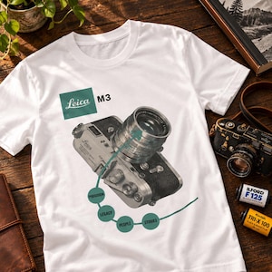 May include: A white t-shirt featuring a graphic of a Leica M3 camera. The design includes a green square with the text Leica M3, and a curved green line connecting four circles labelled Precision, Legacy, People, and Stories in white text.